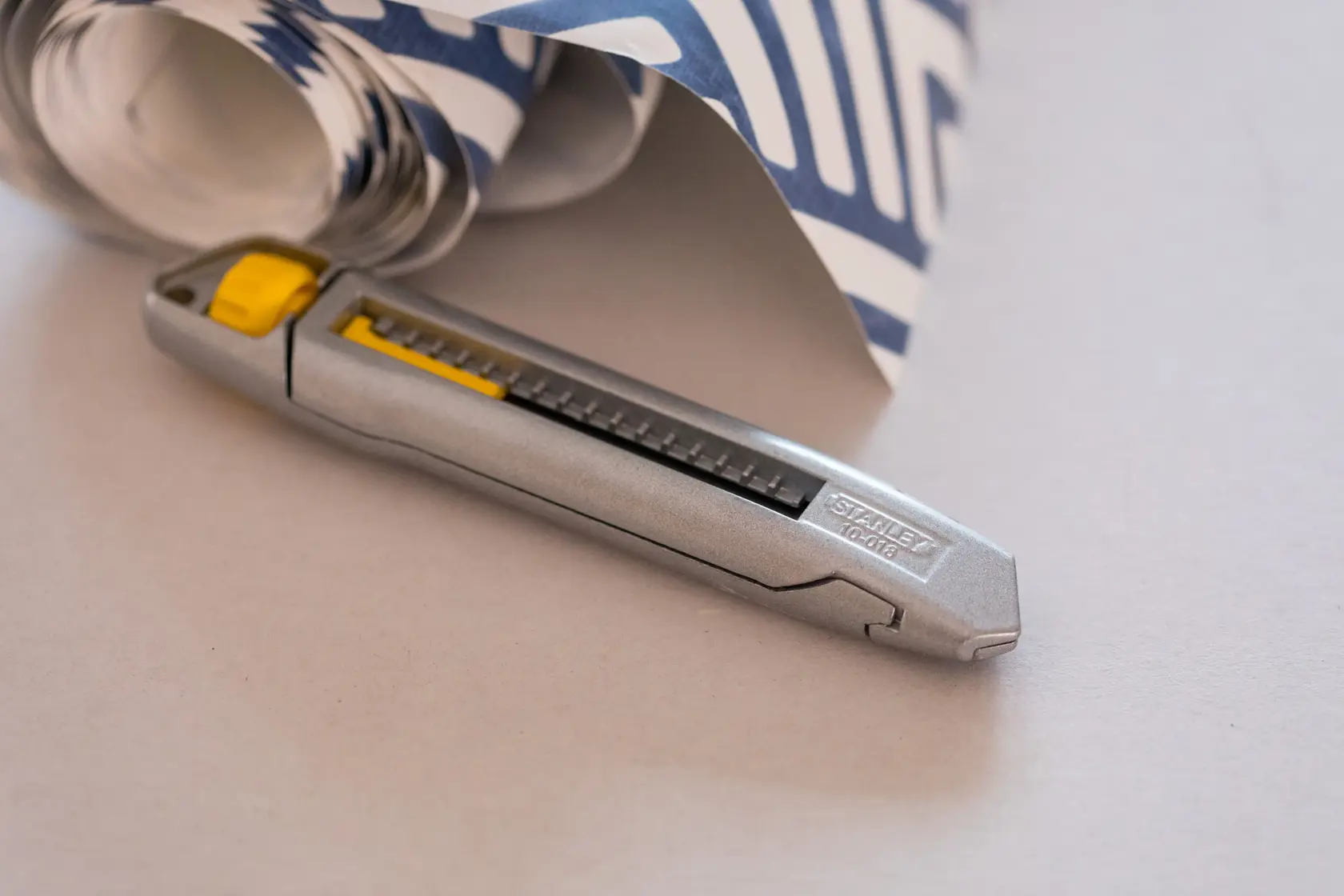 STANLEY 1-10-018 utility knife with silver body and yellow slider, shown laying next to rolled patterned paper.