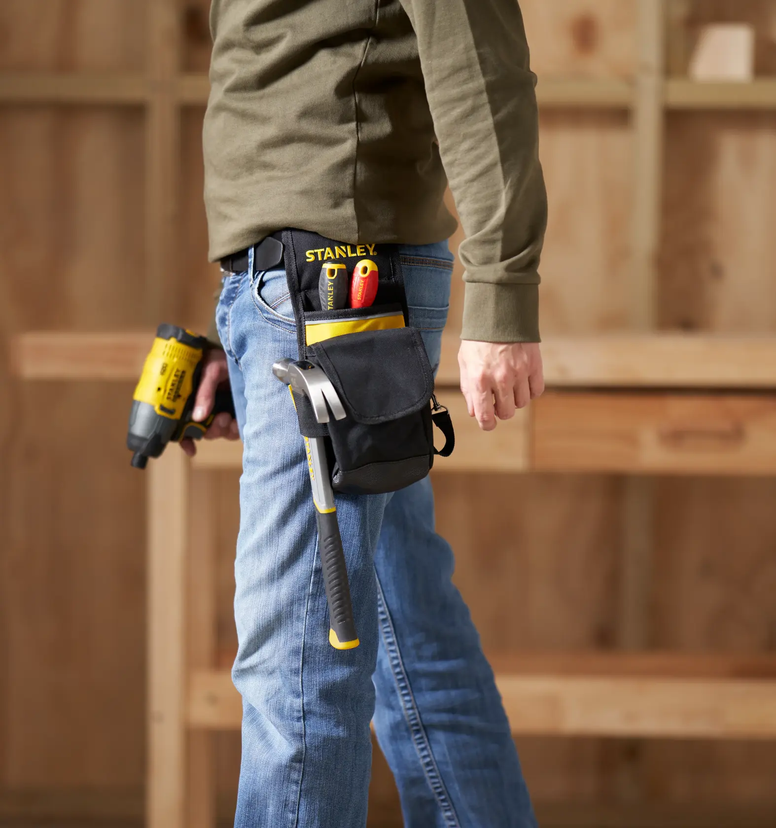 STANLEY 1-93-329 Werkzeugtasche mit Hammer, Schraubendrehern und Universalmesser am Gürtel, Seitenansicht, Werkstatt im Hintergrund.