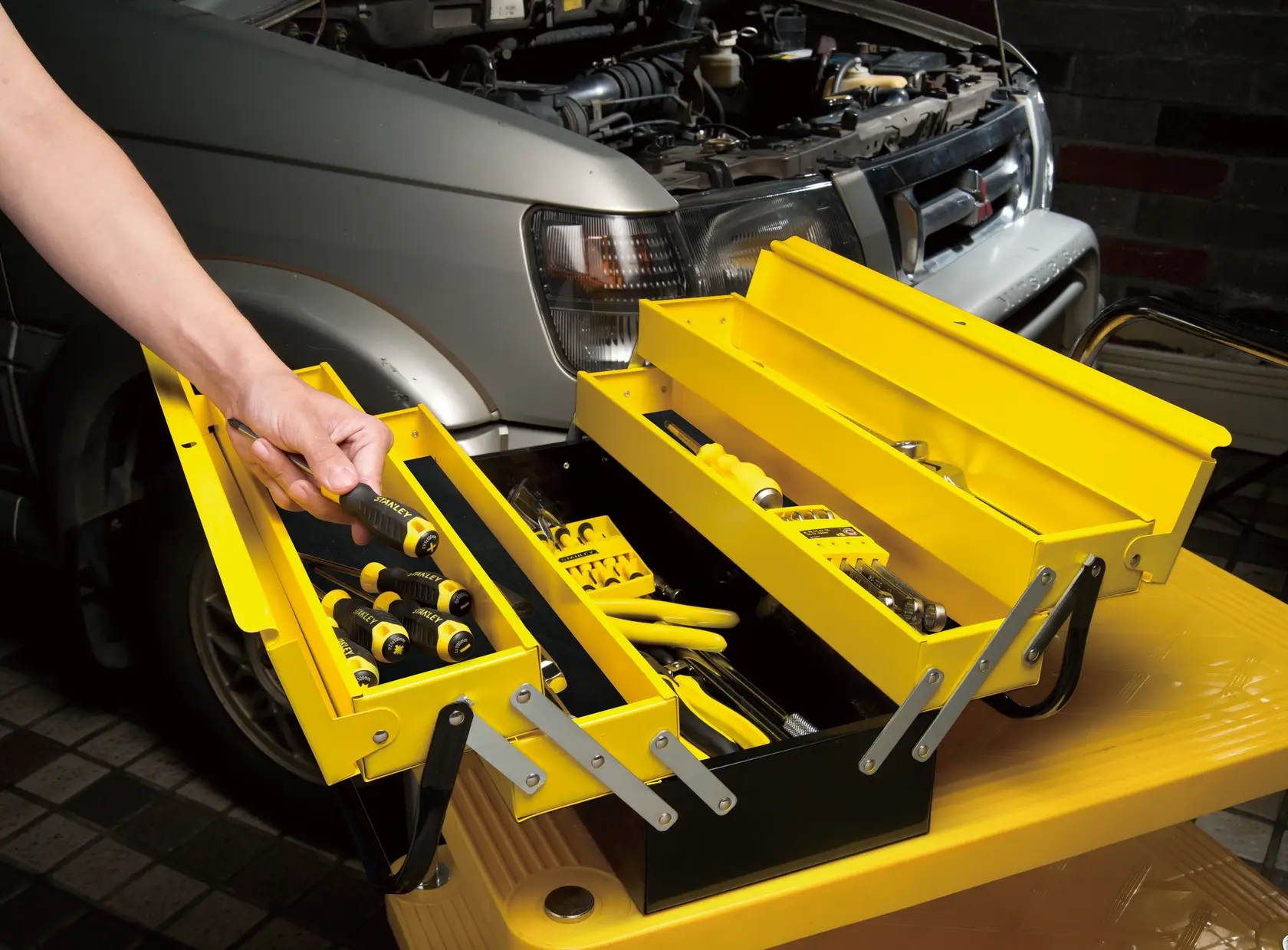 STANLEY 1-94-738 yellow metal toolbox open with tools, shown near a car engine, side view.