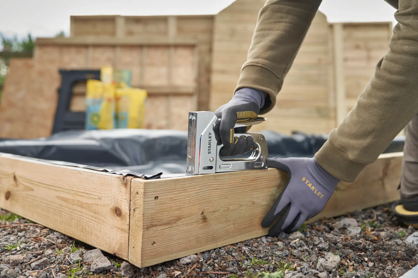 Photo of STANLEY 6-TR110 stapler being used to fasten plastic liner to wooden garden bed frame outdoors.