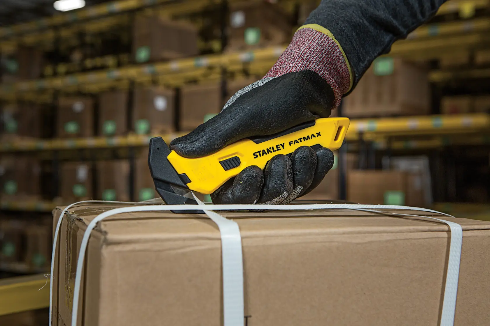 Stanley FMHT10358-0 utility knife held by gloved hand cutting box strap in warehouse.