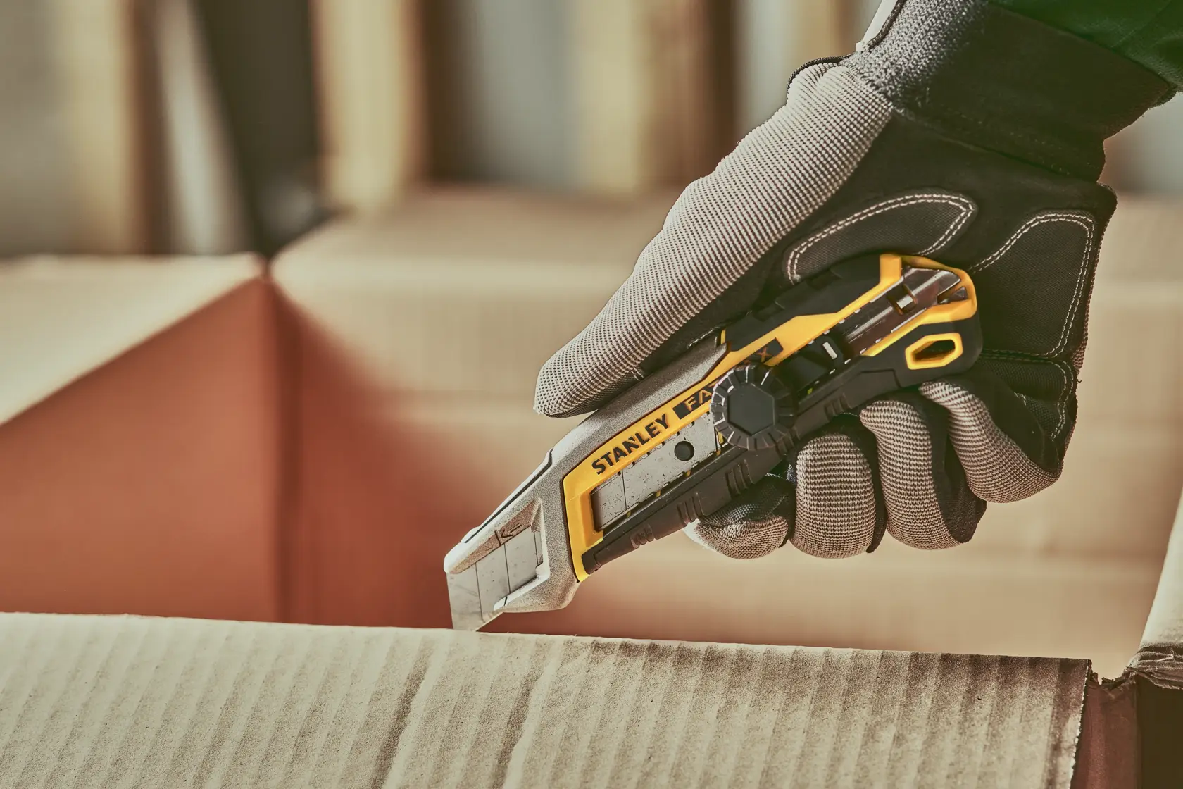 Stanley FMHT10592-0 utility knife cutting cardboard, shown in gloved hand, product photography.