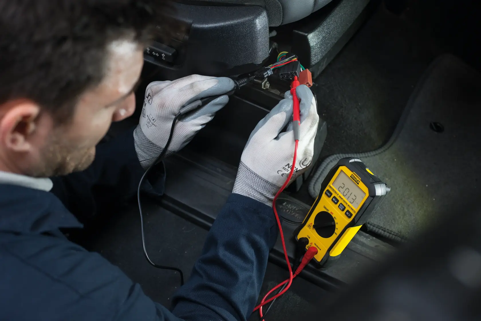 Photo of STANLEY FMHT82563-0 digital multimeter in use testing car wiring, yellow body, clearly visible display.
