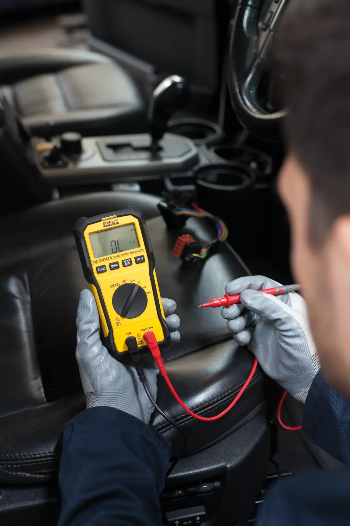 Photo of STANLEY FMHT82563-0 digital multimeter in use inside a car, with gloved hands and test leads.