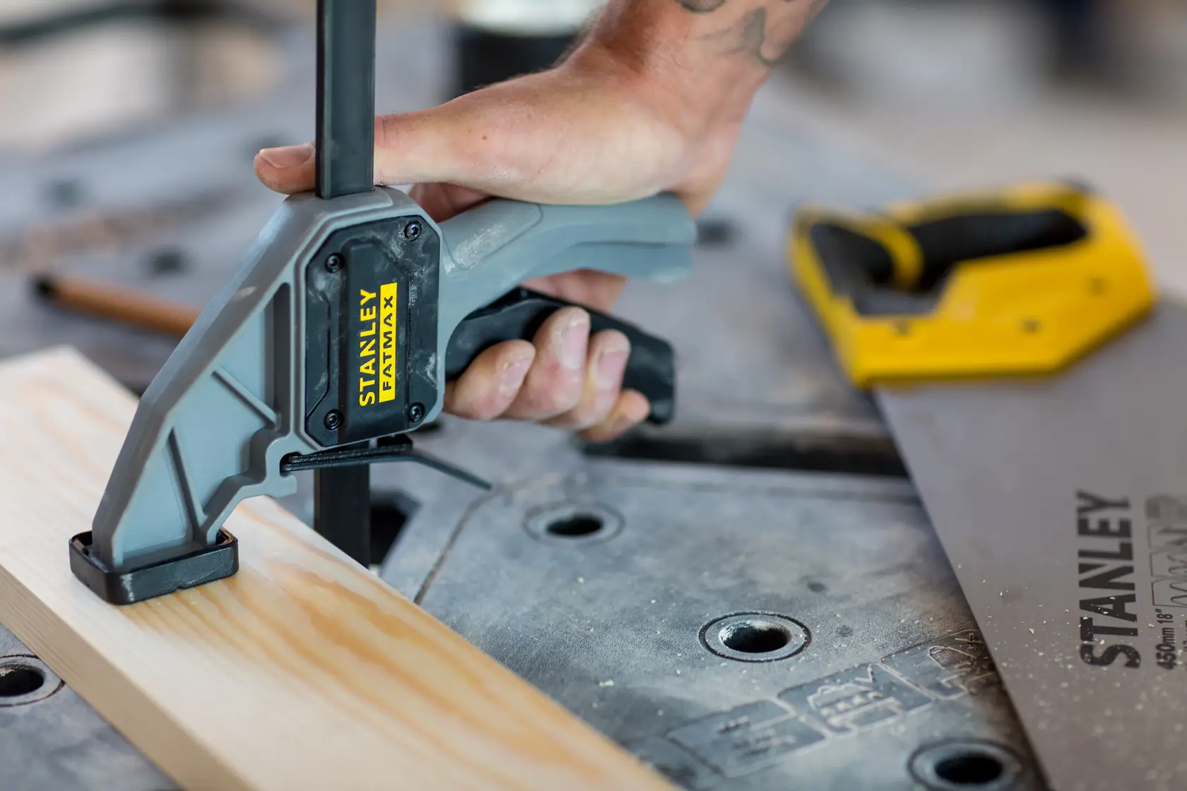 STANLEY FMST1-75672 clamp securing wood on workbench, close-up with hand gripping handle, saw in background.