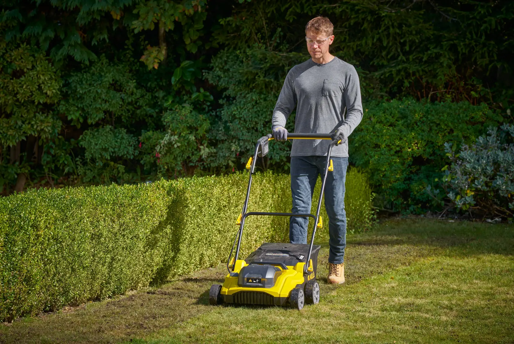 STANLEY SCOEP3801N yellow lawn scarifier being used in a garden, with greenery in the background.