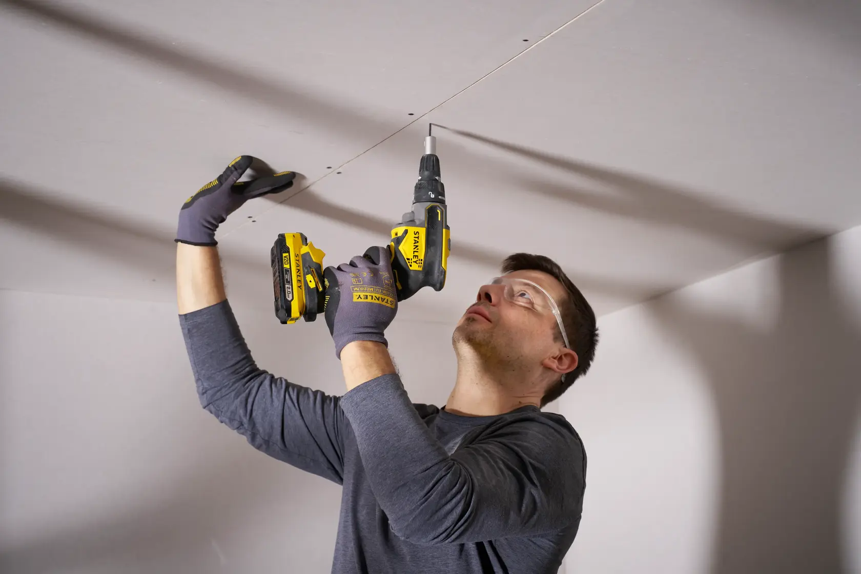 Worker using STANLEY SFMCF600B cordless drill to fasten screws into ceiling drywall, indoor construction scene.