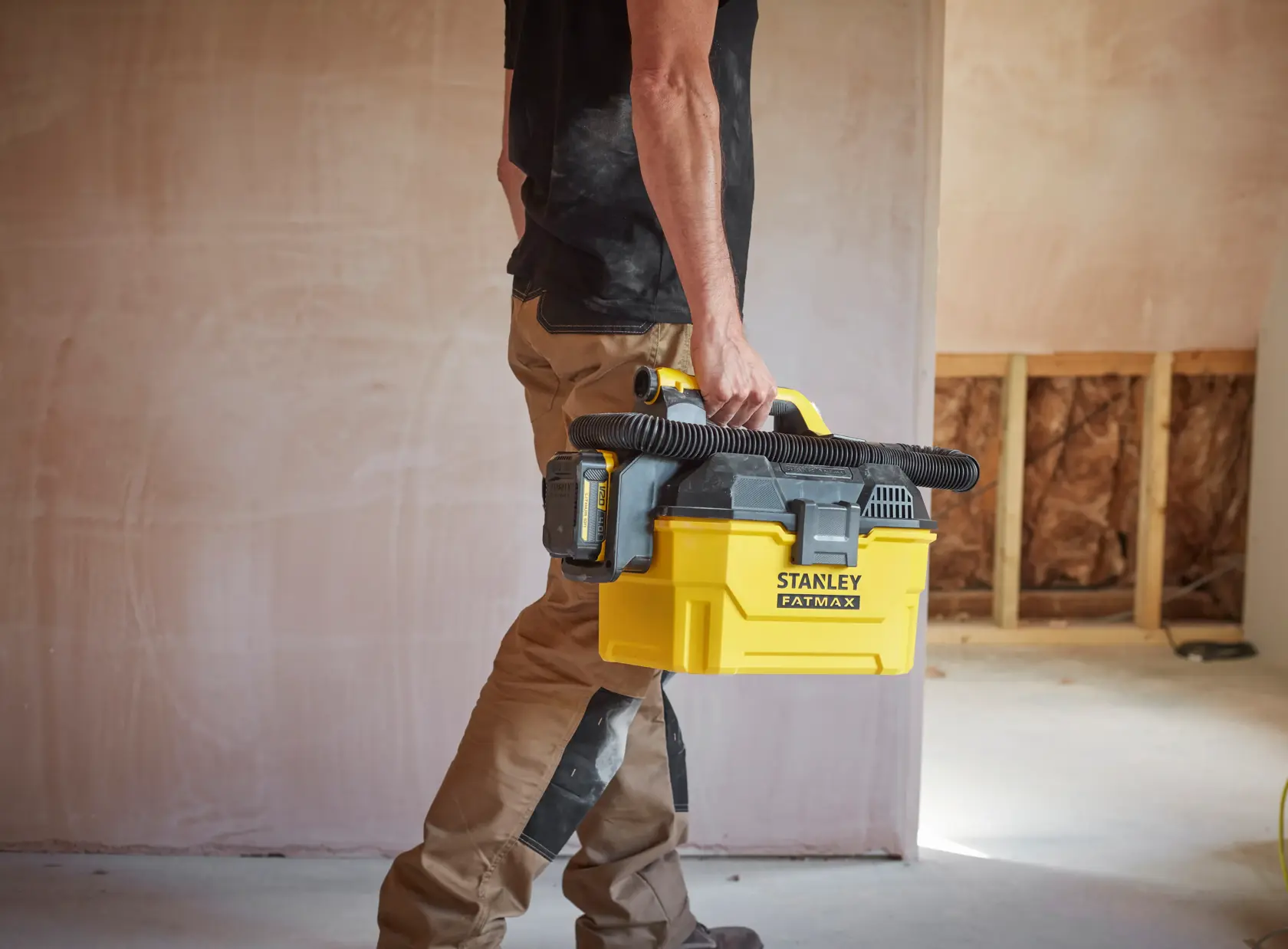 Person trägt STANLEY SFMCV002B tragbaren gelben Staubsauger mit schwarzem Schlauch auf einer Baustelle.