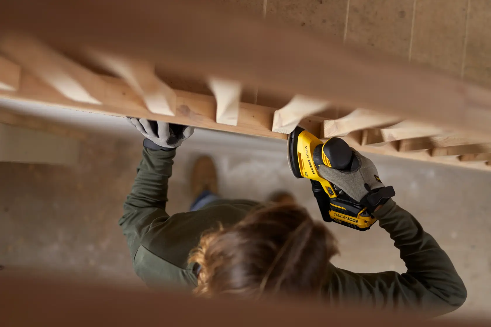 Foto eines Arbeiters mit STANLEY SFMCW221B Schleifer an einer Holztreppe, mit Handschuhen, Innenbereich.