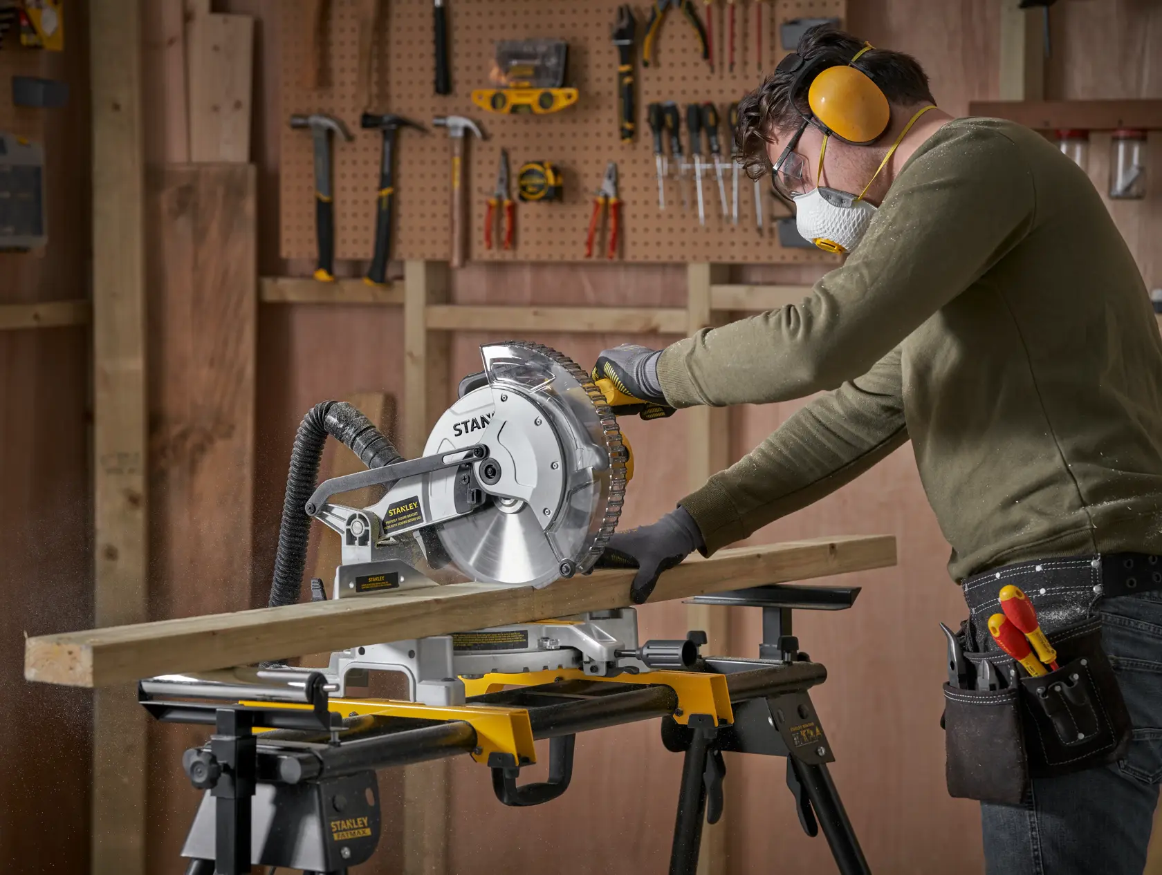 Photo of a person using the STANLEY SM1650 miter saw to cut wood, in a workshop with tools on a pegboard wall.