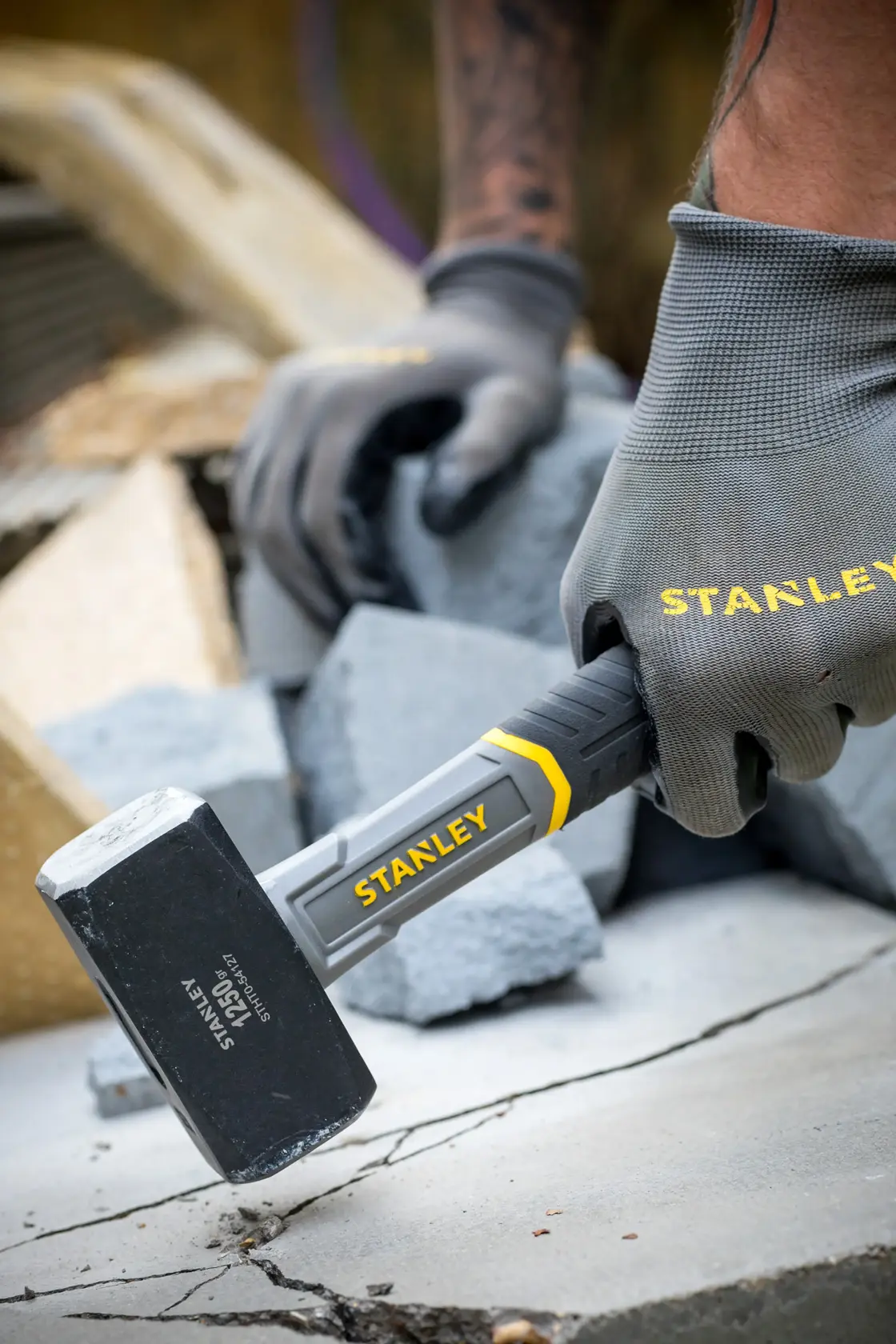Photo of STANLEY STHT0-54127 club hammer in use, black head with yellow accents, held by gloved hand on concrete.