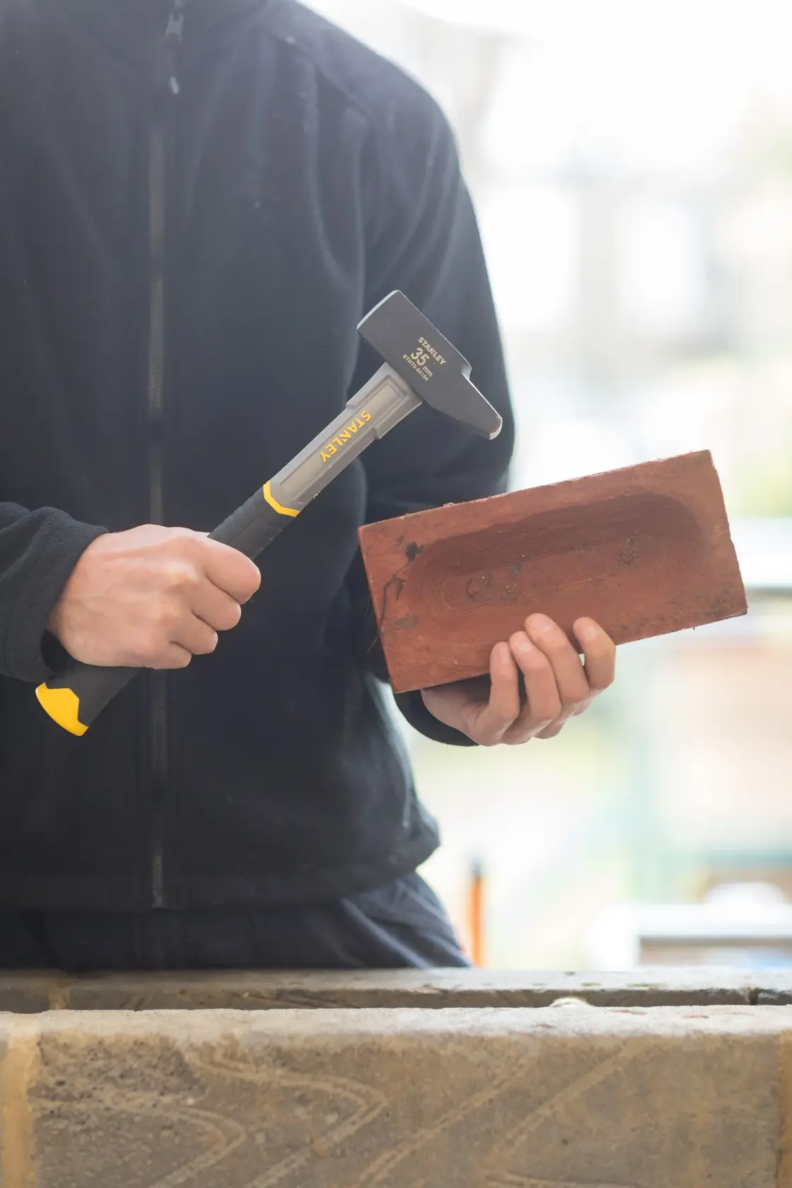STANLEY STHT0-54154 hammer with black and yellow handle held by person in black jacket, used on red brick.