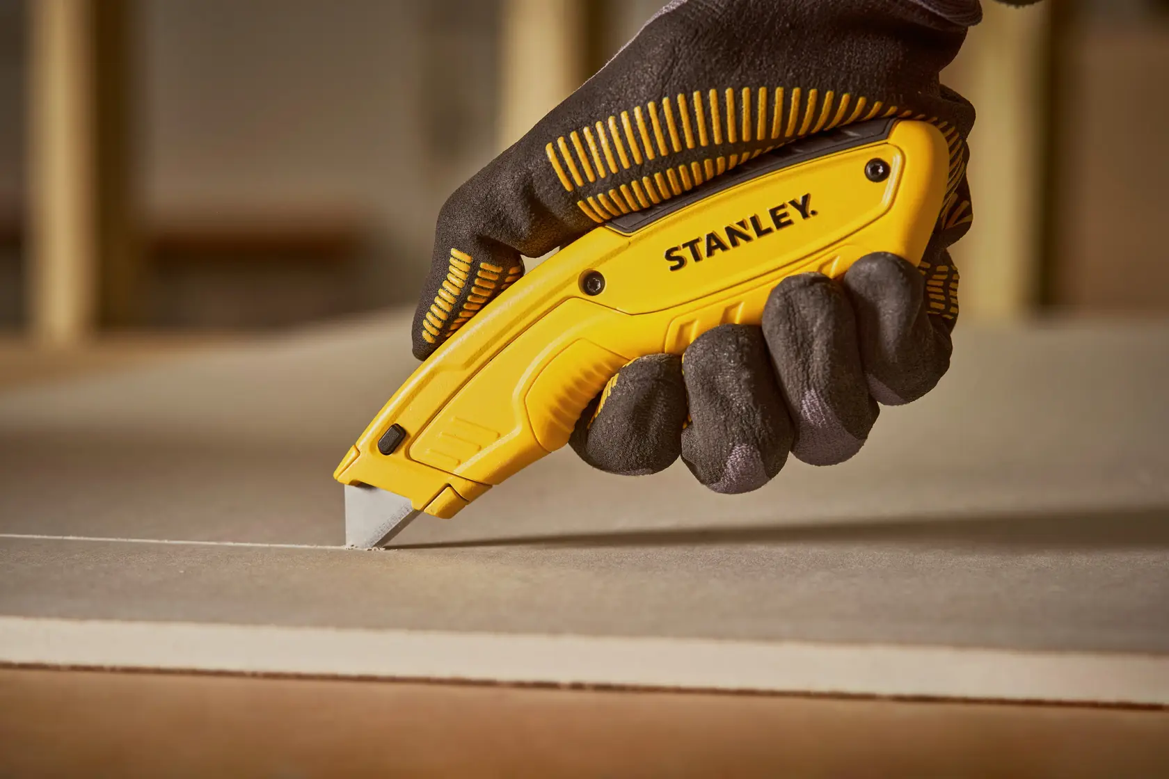 Photo of STANLEY STHT10430-0 yellow utility knife in gloved hand cutting drywall, side view.