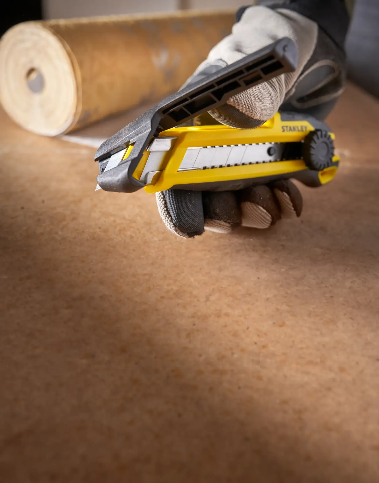 STANLEY STHT10500-0 utility knife held by gloved hand on work surface, roll in background, close-up angle.