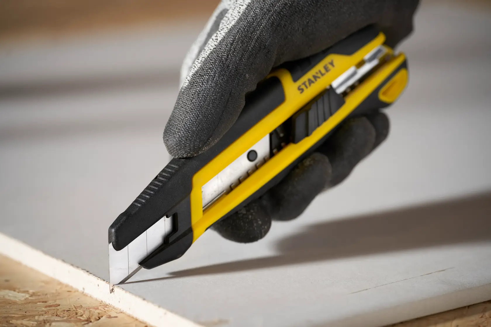 Photo of STANLEY STHT10500-0 utility knife cutting drywall, black and yellow handle, hand in glove holding tool.