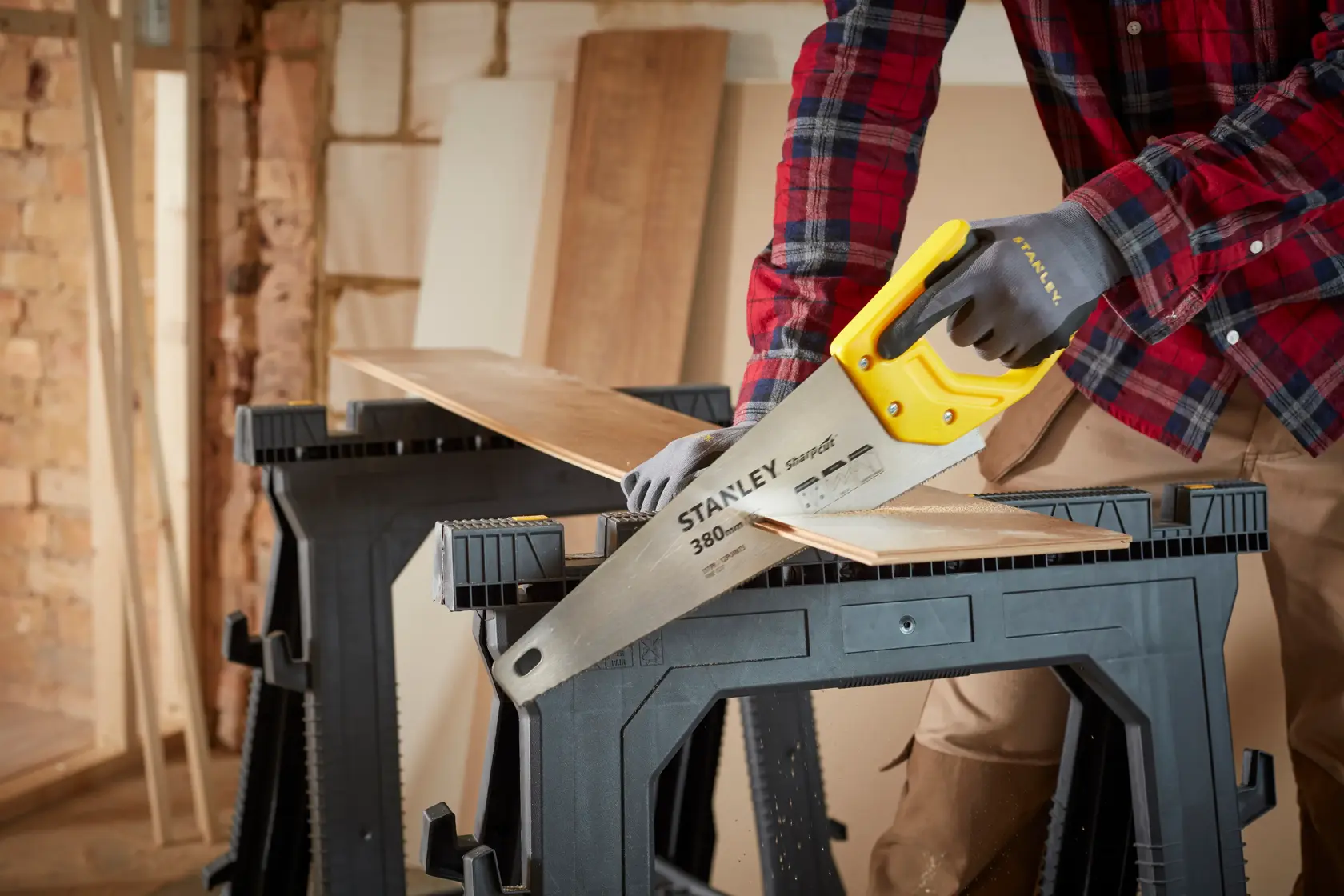 Stanley STHT20369-1 hand saw with yellow handle cutting wood plank on black sawhorses in workshop.