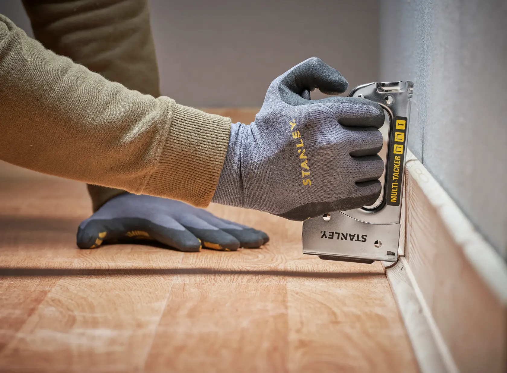 STANLEY STHT6-70410 Multi-Tacker used for flooring, close-up of gloved hand stapling near wall.