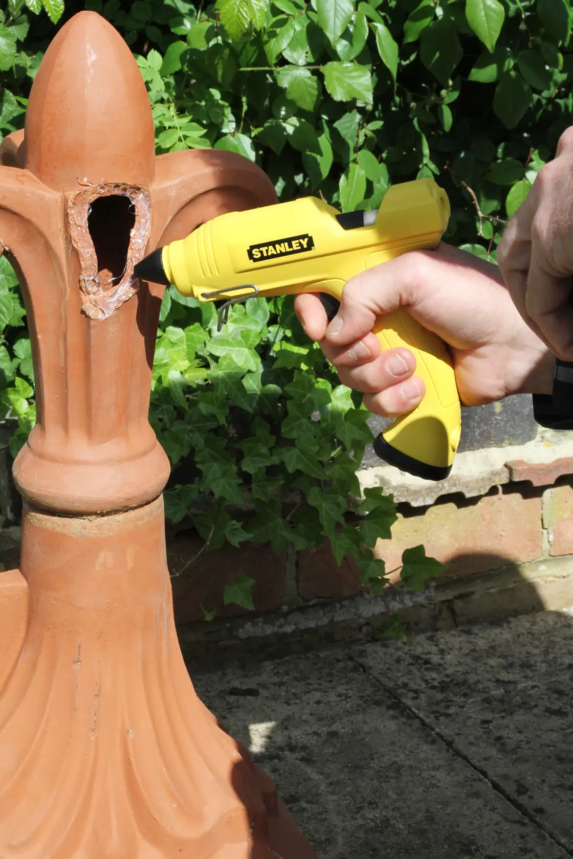 Stanley STHT6-70416 yellow glue gun used on outdoor ceramic ornament, close-up, garden background.