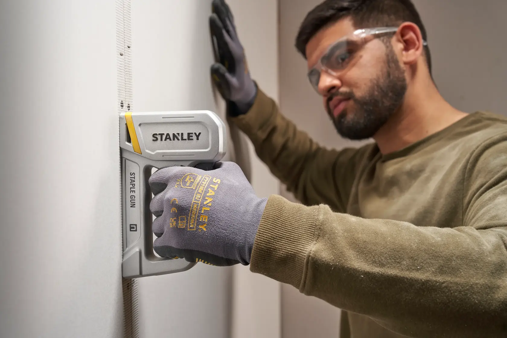 Stanley STHT70150-0 staple gun in use on drywall by gloved hand, close-up side angle, photography product.