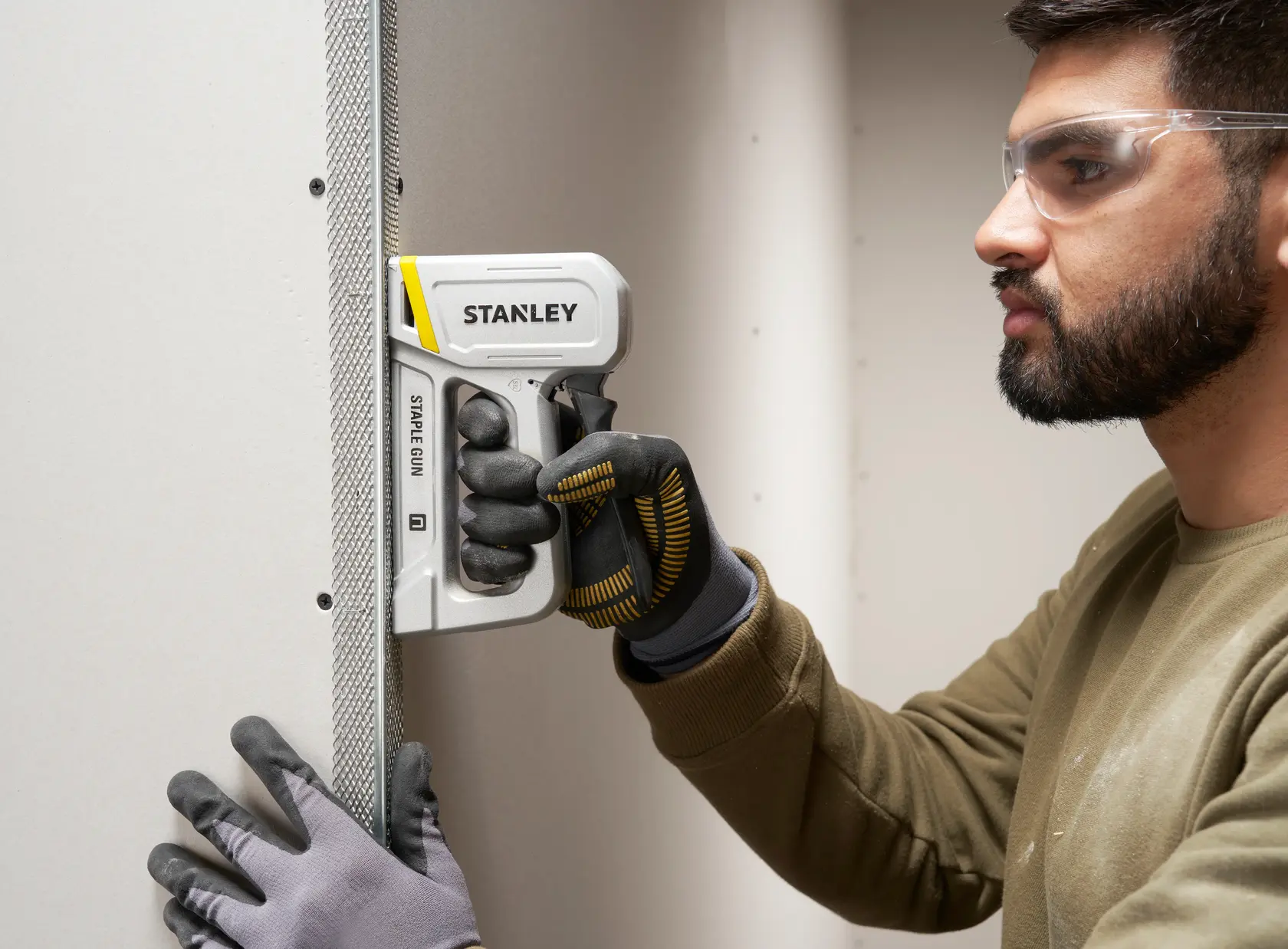 Stanley STHT70150-0 staple gun held by a person in gloves, used to attach metal corner bead to drywall.