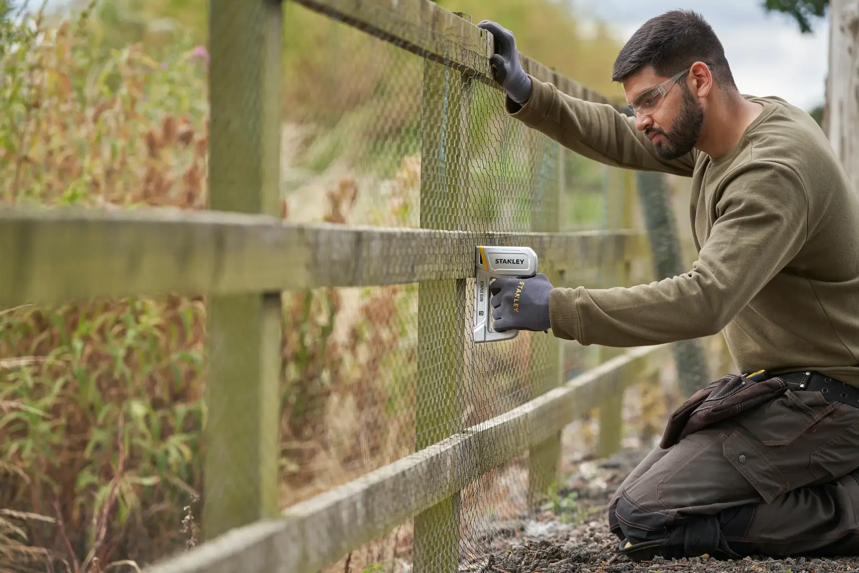 Photo of a person using a STANLEY STHT70150-0 stapler to attach wire mesh to a wooden fence outdoors.