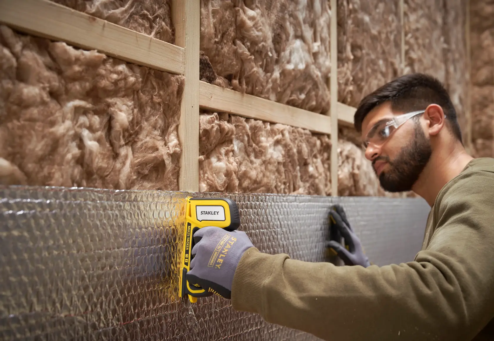 STANLEY STHT70440-0 stapler in use attaching foil insulation to wall, gloved hand visible, side angle.