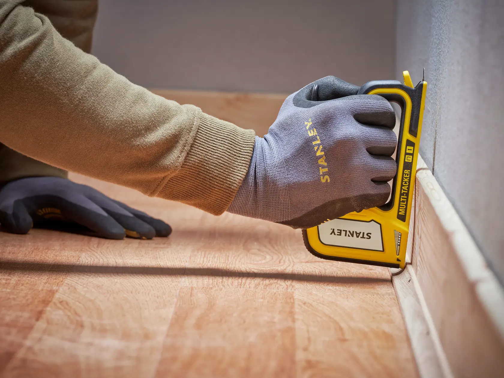 Photo of STANLEY STHT70440-0 stapler in use, gloved hand securing baseboard to wall, close side view.