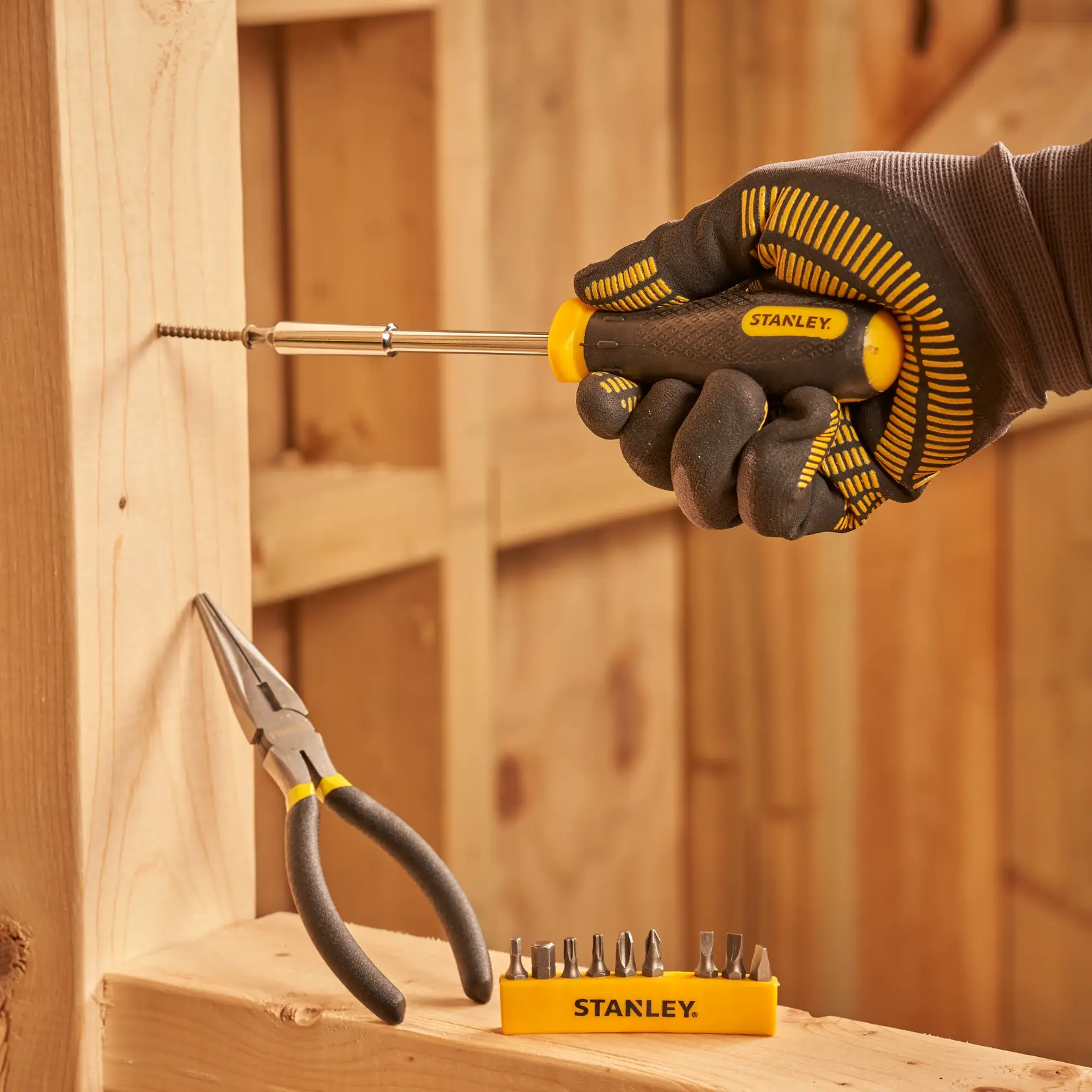 STANLEY STMT0-74101 screwdriver set with bits, gloved hand using tool, pliers nearby, indoor woodworking.