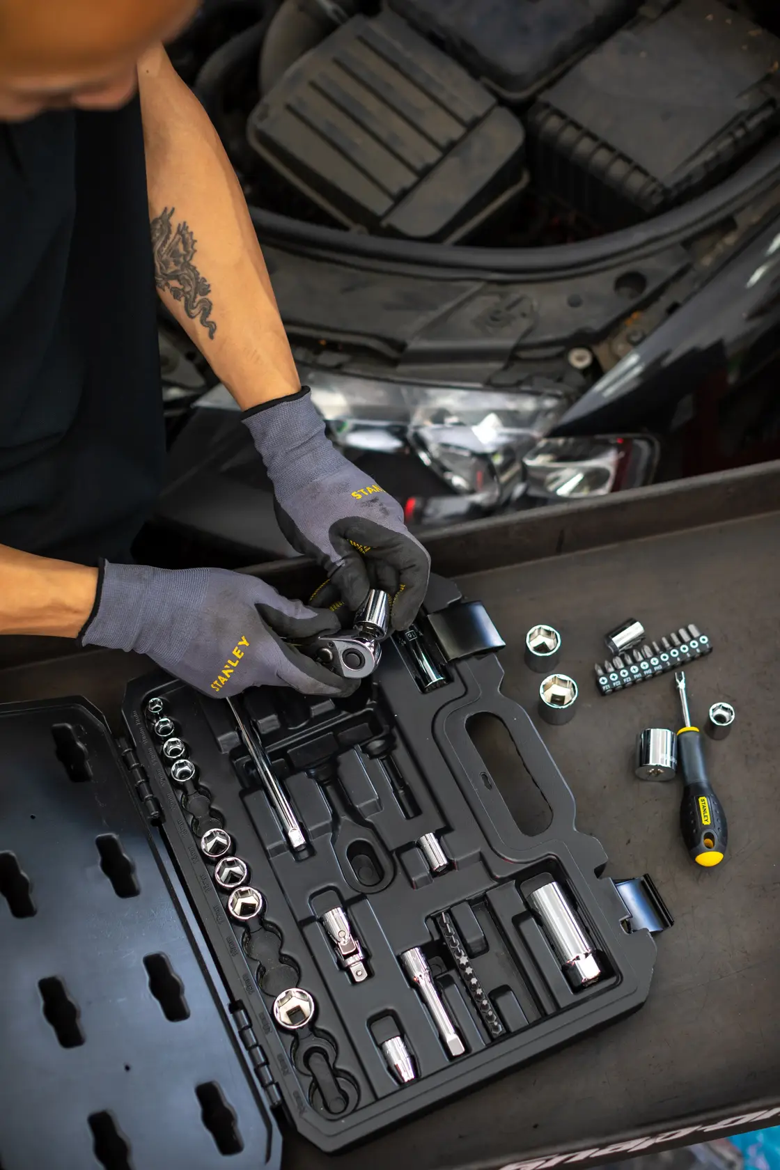 STANLEY STMT82828-1 socket set in black case, mechanic using tools near car engine, photography from above.