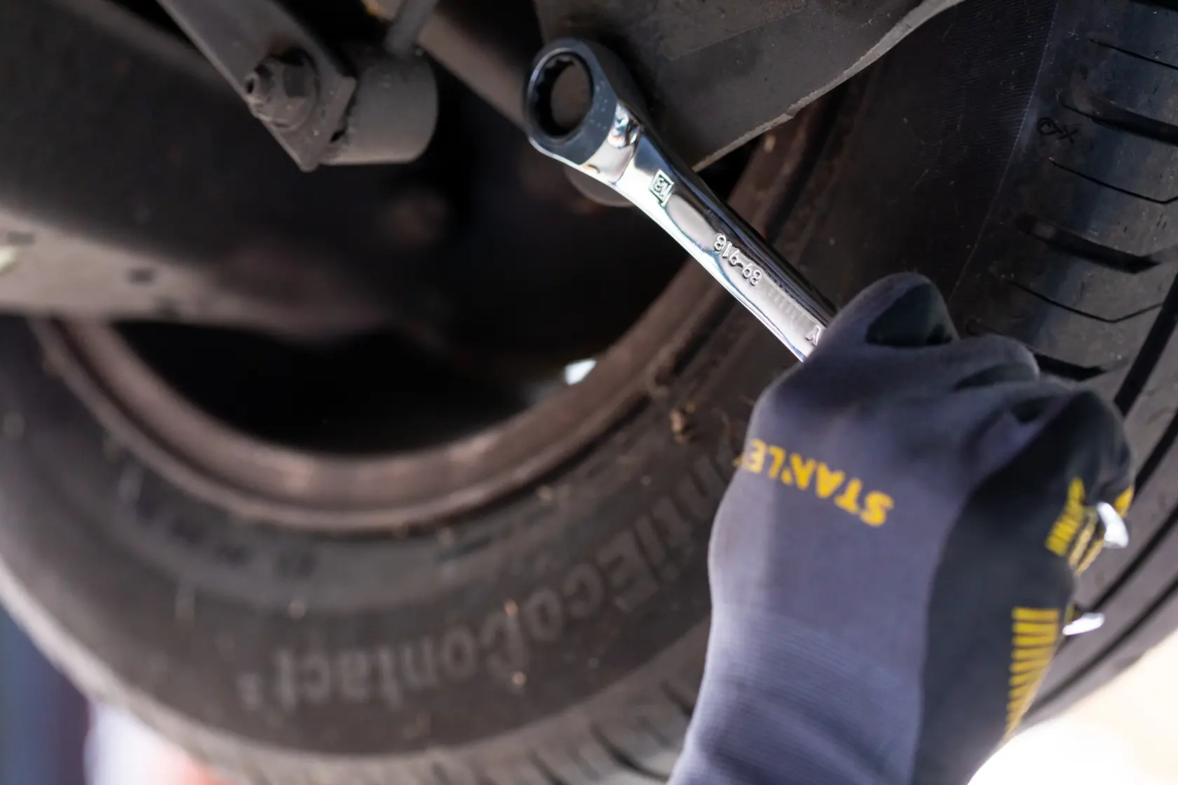 Photo of STANLEY STMT89917-0 ratcheting wrench used on car wheel, gloved hand visible, automotive context.