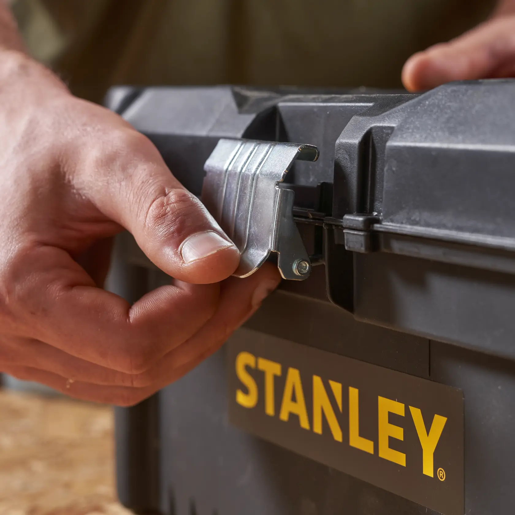 STANLEY STST1-75518-KR Werkzeugkasten Nahaufnahme, Hand öffnet Metallschließe, schwarzes Gehäuse, gelbes Logo.