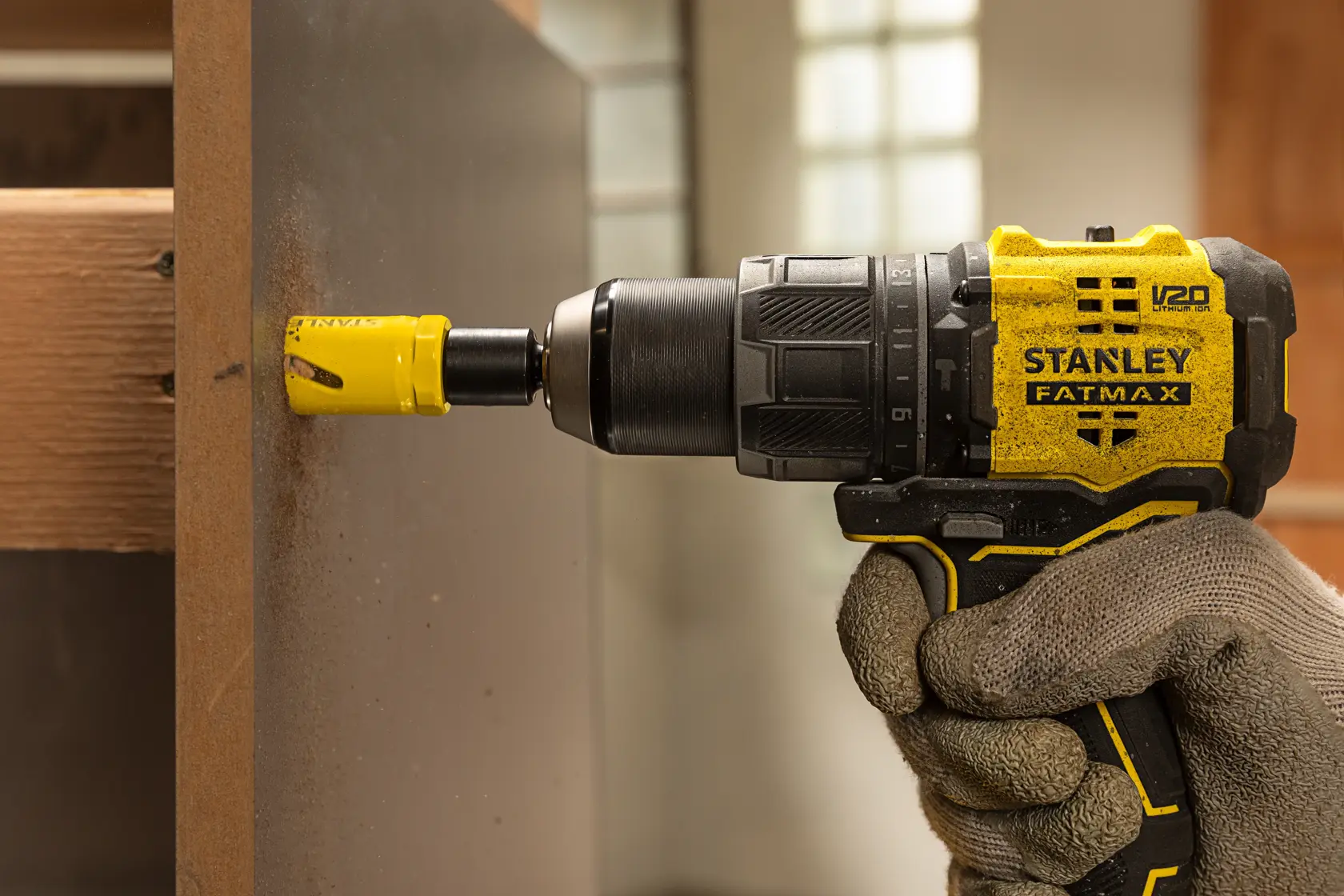 A gloved hand holds a yellow and black Stanley Fatmax power drill with a hole saw attachment, drilling through a wooden panel indoors.